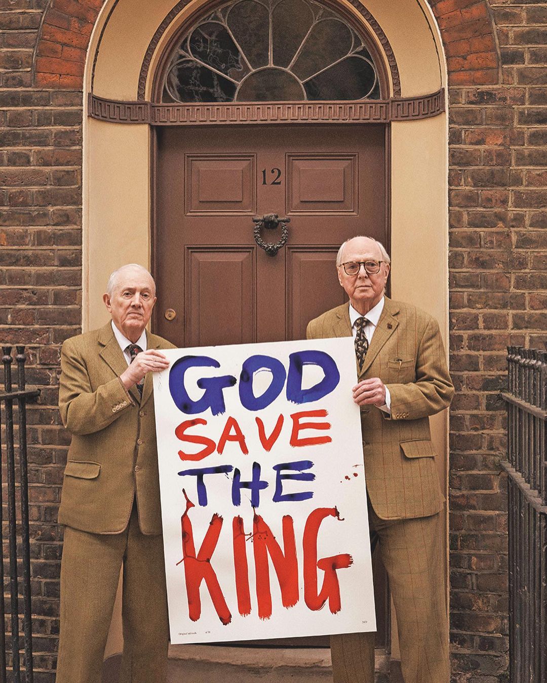 Two elderly men in tweed suits stand before a brown door, holding a sign reading 'God Save the King' in bold red and blue letters.
