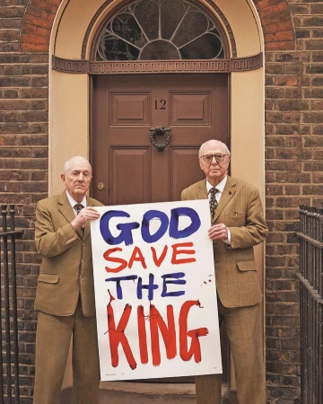 Two elderly men in tweed suits stand before a brown door, holding a sign reading 'God Save the King' in bold red and blue letters.