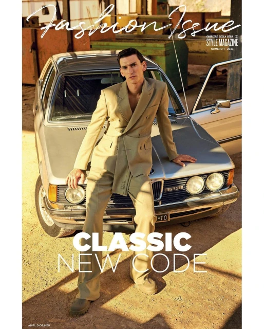 Fashionable man in suit poses with vintage silver car for magazine cover, standing outdoors in earth-tone environment with natural lighting.