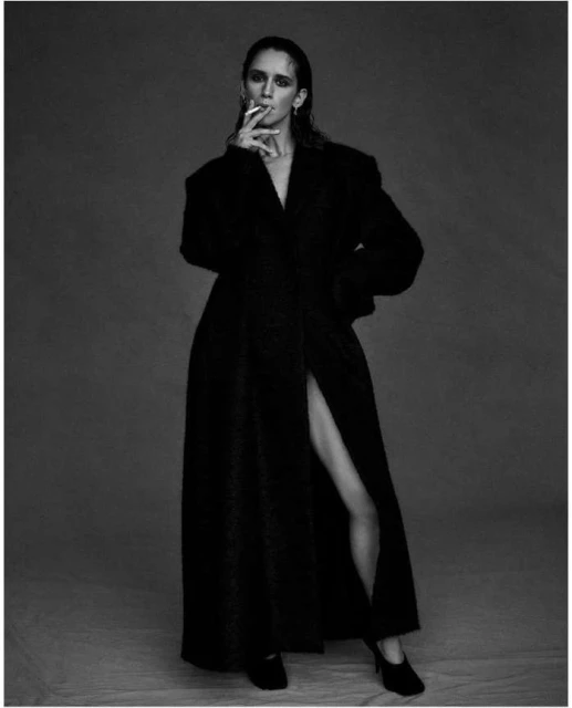 Woman in a long coat and heels, holding a cigarette, standing indoors against a plain grey background in a black and white portrait.