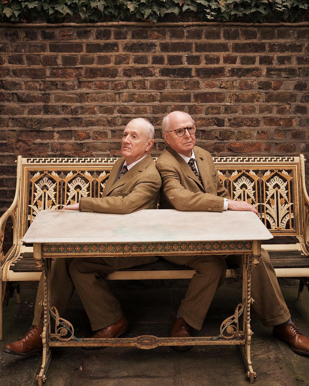Two elderly men in suits sit back-to-back at a table on a bench against a brick wall with ivy, in an outdoor setting.