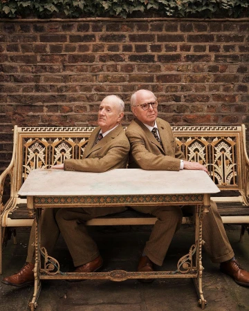 Two elderly men in suits sit back-to-back at a table on a bench against a brick wall with ivy, in an outdoor setting.