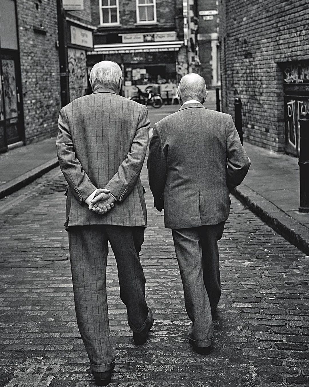 Black and white photo of two elderly men walking down a cobblestone street lined with brick buildings in an urban setting.