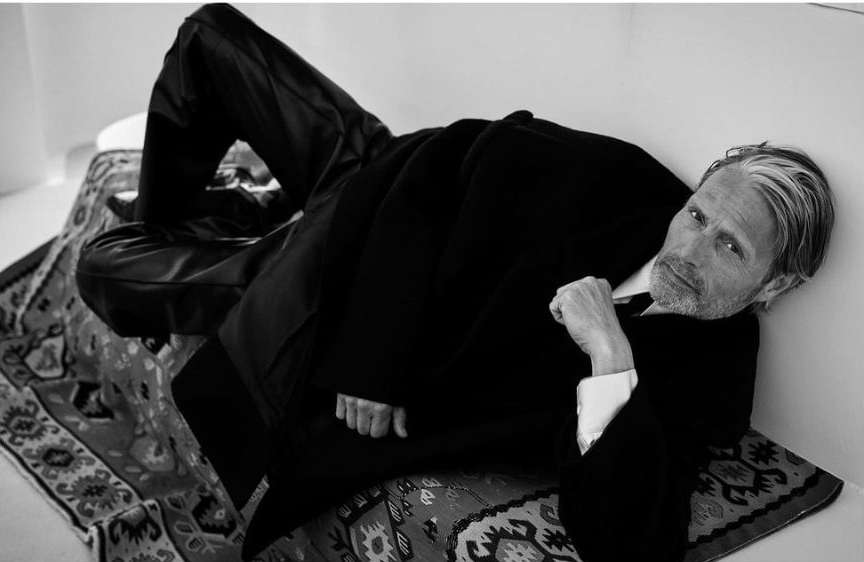 Monochrome portrait of an adult male lying on a patterned cushion, wearing a dark jacket and trousers, looking toward the camera.