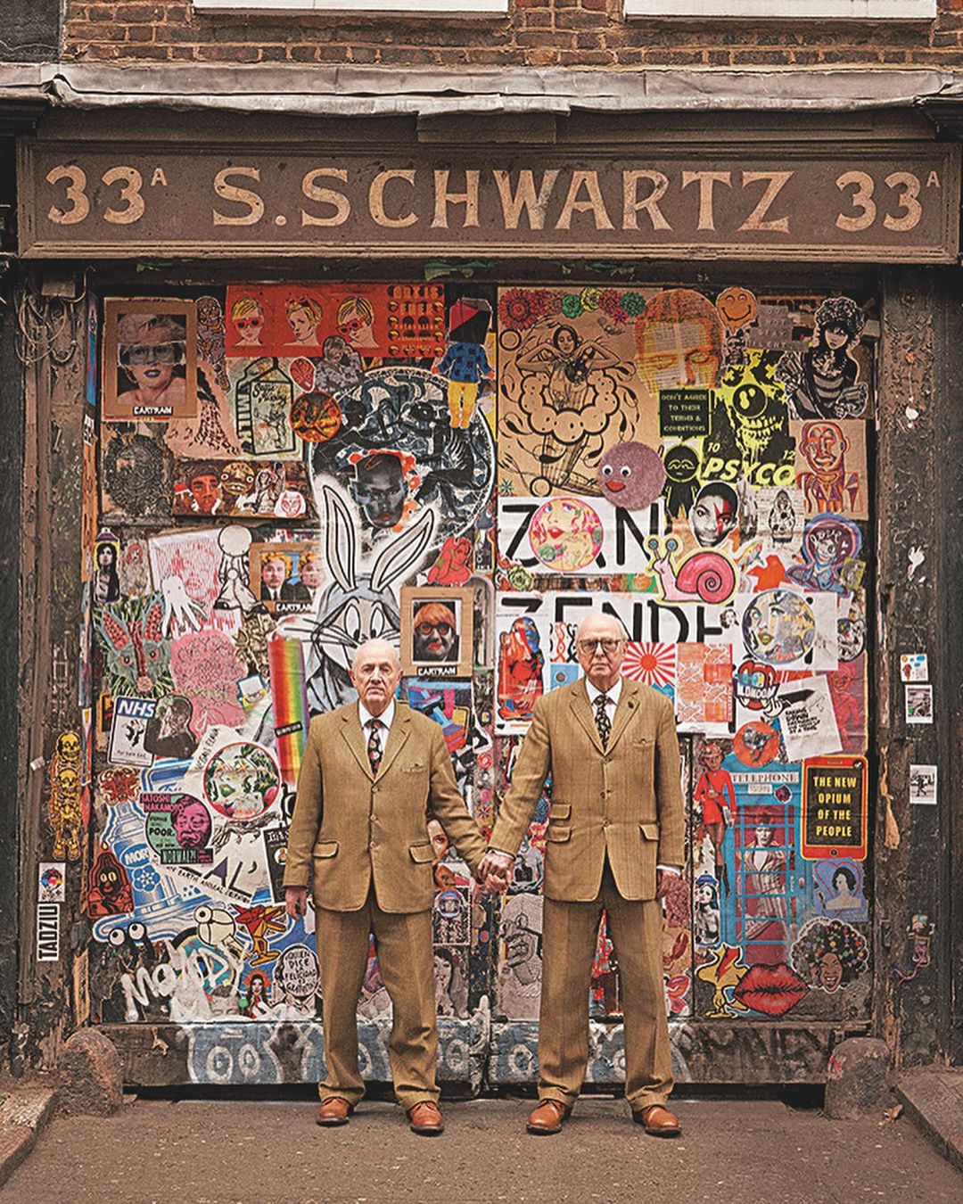 Two elderly adult males pose in matching tweed suits, standing in front of a weathered, sticker-covered brick doorway, vintage style.