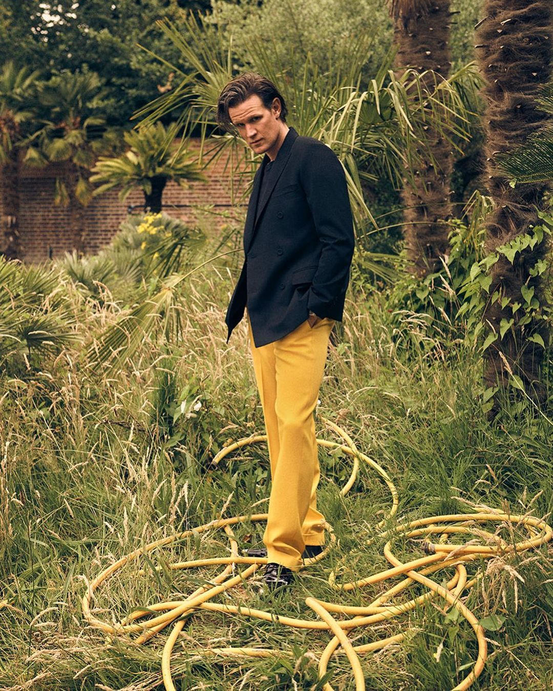 A man in a dark jacket and yellow trousers stands in a grassy garden with palm trees, posing for a portrait shot in natural lighting.