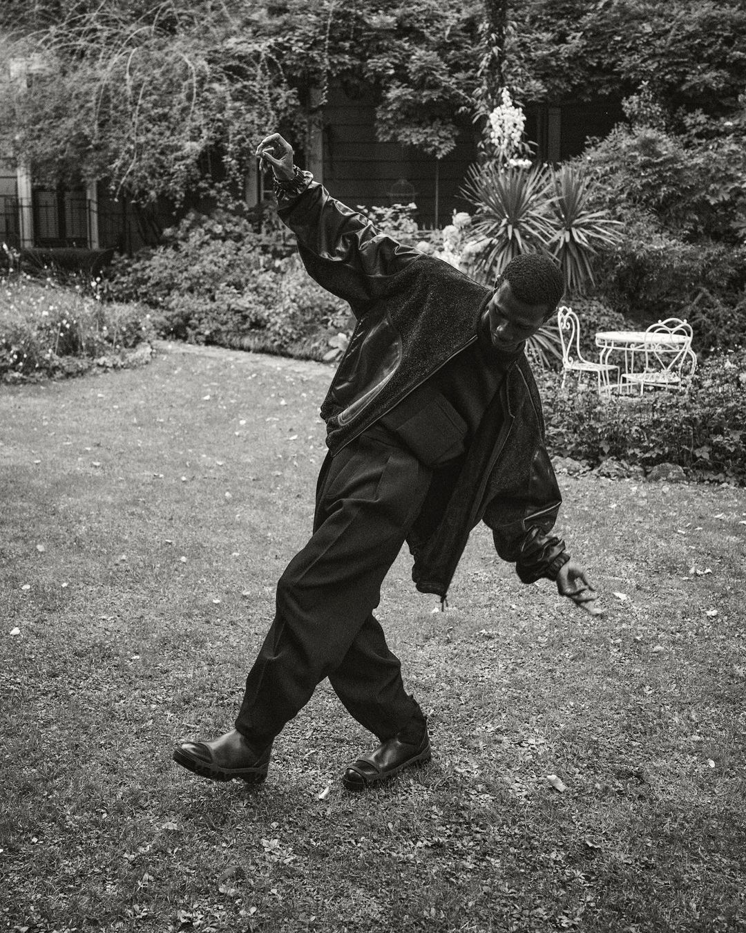 Monochrome portrait of an African-American adult male posing with outstretched arms in a garden setting with plants and white furniture.