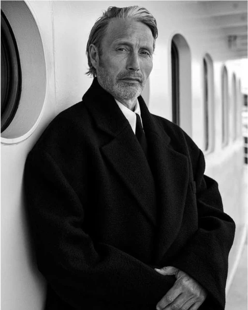 Mads Mikkelsen poses in a black coat against a white wall with porthole windows in a black and white portrait, exuding understated elegance.
