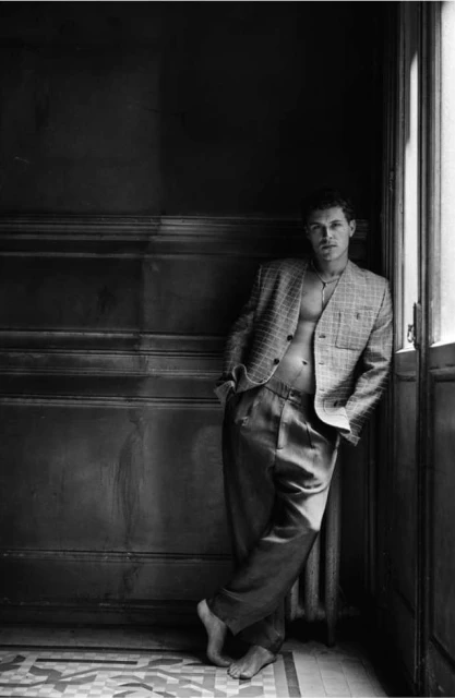 Black and white studio portrait of a man in an open blazer and pants, leaning against a wall with a patterned floor. 