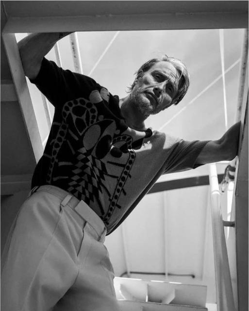 Monochrome portrait of a man with short hair, in a printed knit shirt and light pants, posing on a staircase with natural lighting.