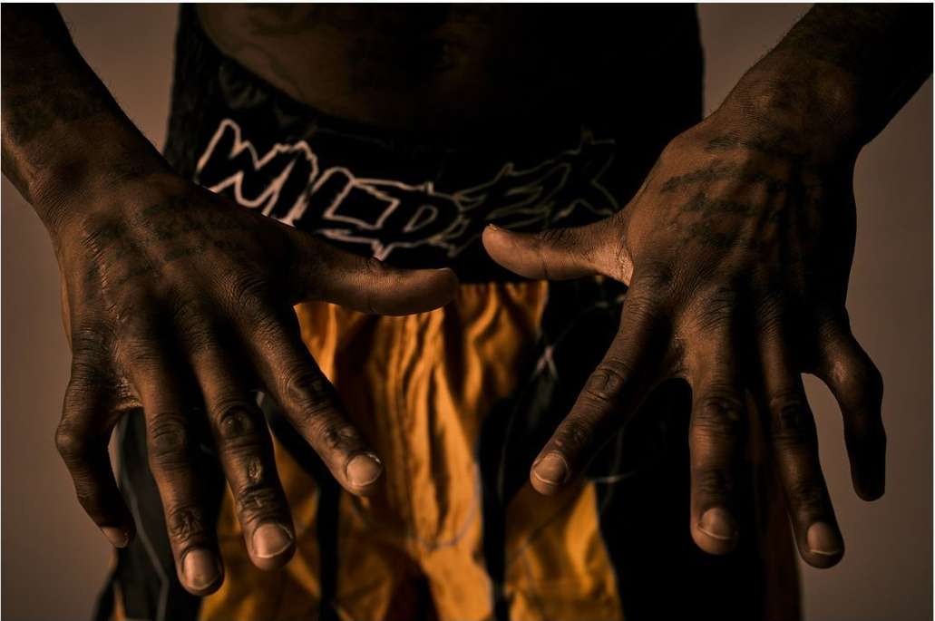 Close-up shot of a person's dark-skinned hands and forearms, showing the texture and musculature, posed against a neutral background.