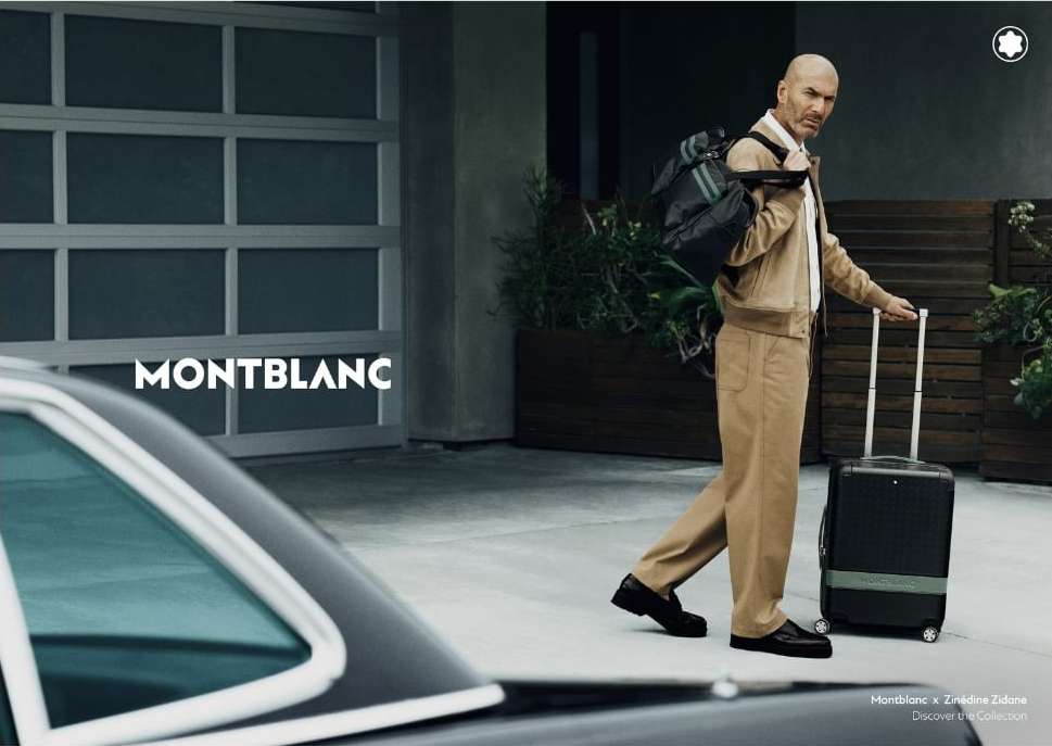 Man in khaki suit walks with suitcase and duffel bag outdoors near a modern car and garage door, advertising Montblanc.