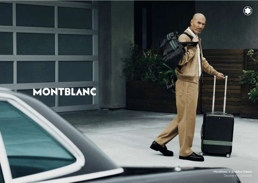 Man in khaki suit walks with suitcase and duffel bag outdoors near a modern car and garage door, advertising Montblanc.