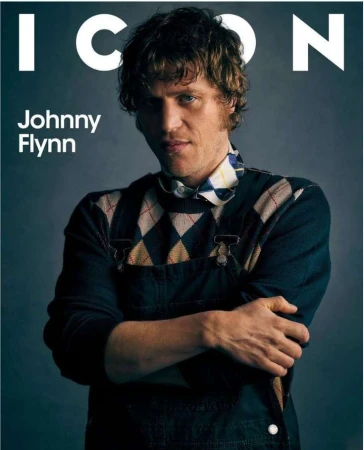 Magazine cover featuring Johnny Flynn wearing a patterned sweater and overalls with his arms crossed against a grey background.