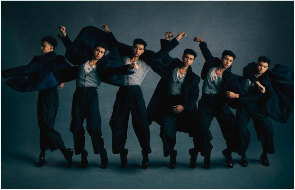 Multiple exposures of a young adult man in a gray vest, black trousers, and a dark coat in a dance-like pose against a gray background.