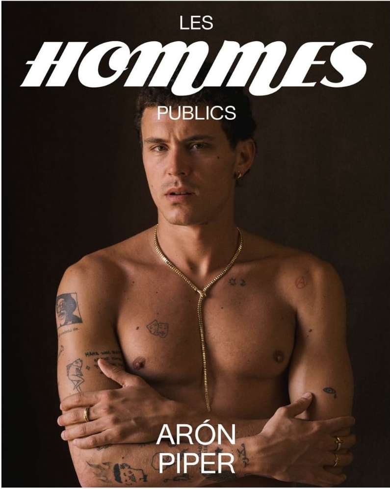 Aron Piper shirtless portrait against dark backdrop, face forward, featuring a gold chain, tattoos, magazine cover, and studio lighting.