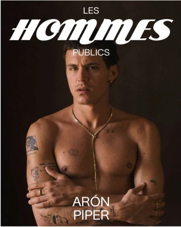 Aron Piper shirtless portrait against dark backdrop, face forward, featuring a gold chain, tattoos, magazine cover, and studio lighting.