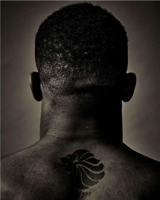 Back view of a dark-skinned person with a closely cropped hairstyle and a lion tattoo on their upper back against a neutral backdrop.