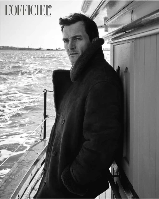 Black and white portrait of a man in a dark coat leaning against the side of a boat with water and shoreline in the distance.