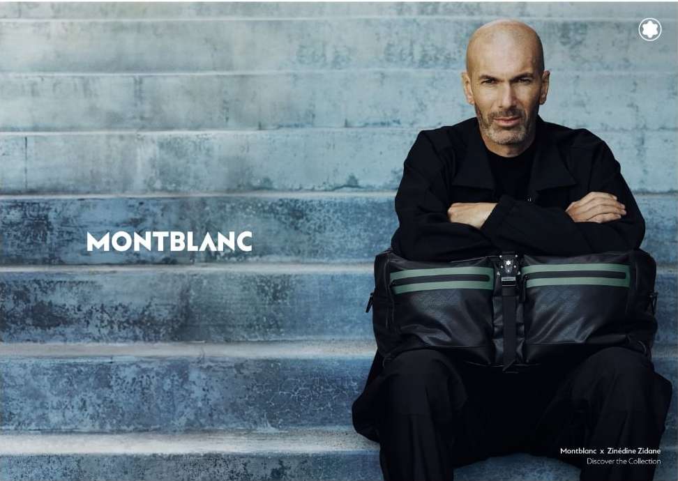 Zinedine Zidane sitting on stairs with a Montblanc bag. He wears a black jacket, and the backdrop features concrete steps in muted blue tones.