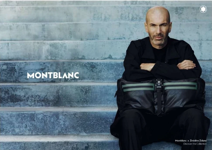 Zinedine Zidane sitting on stairs with a Montblanc bag. He wears a black jacket, and the backdrop features concrete steps in muted blue tones.