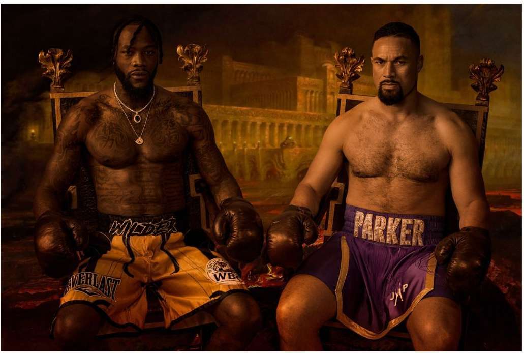 Two shirtless male boxers, Deontay Wilder and Joseph Parker, sit on thrones with boxing gloves, posing in an indoor environment with a backdrop of coliseum.