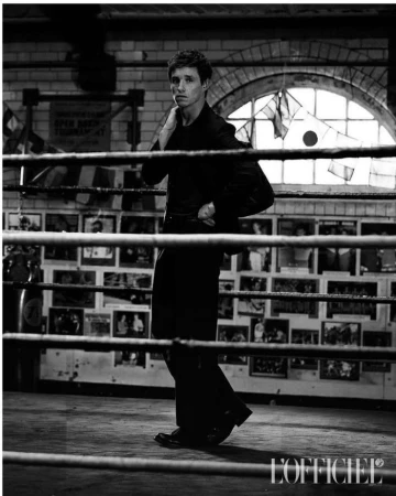 Eddie Redmayne in a dark suit standing inside a boxing ring with a tiled wall background by photographer Charlie Gray