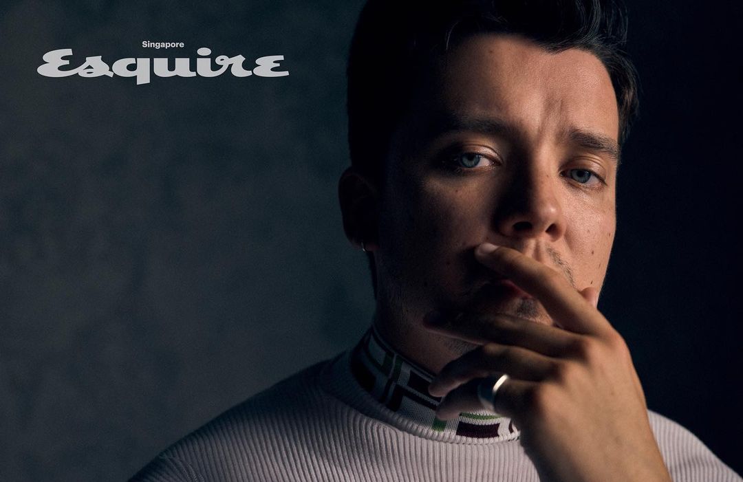 Close-up portrait of a man with blue eyes and dark hair, wearing a white turtleneck against a dark grey background in soft lighting.