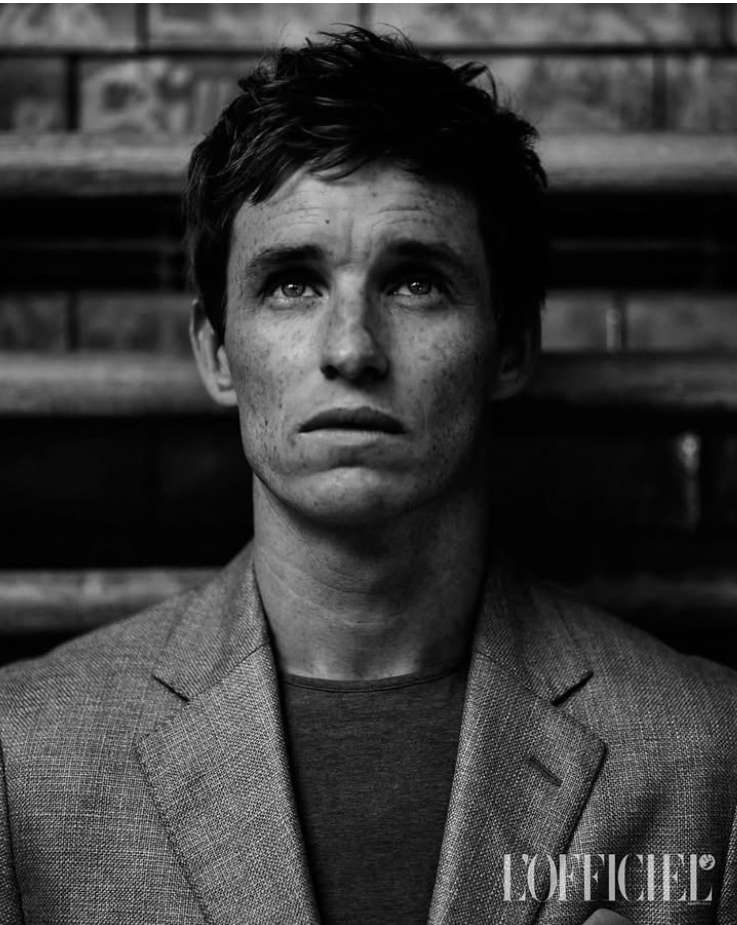 Black-and-white portrait of fair-skinned adult male in suit jacket against blurred brick background, looking upwards with neutral expression.
