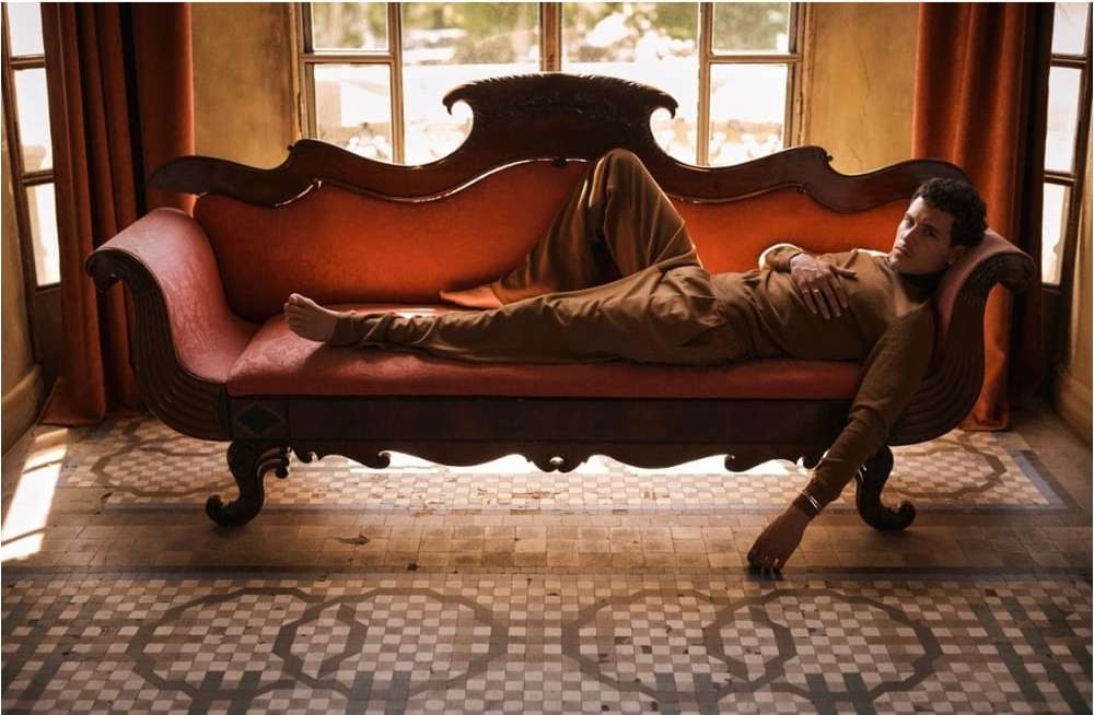 Adult male in brown loungewear reclines on an ornate red chaise lounge indoors with patterned tile flooring and natural window light.