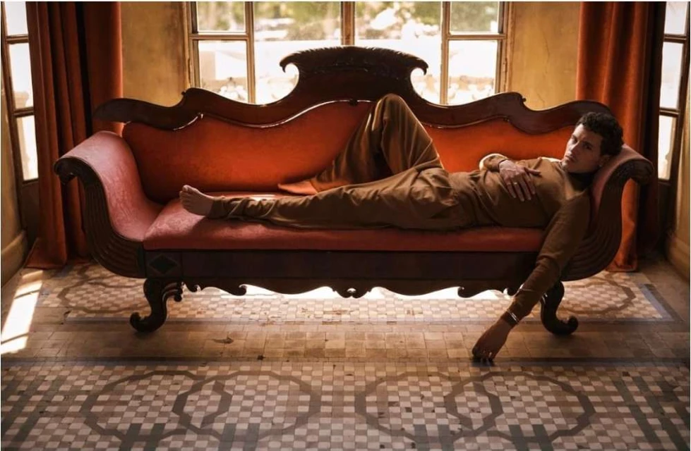 Adult male in brown loungewear reclines on an ornate red chaise lounge indoors with patterned tile flooring and natural window light.