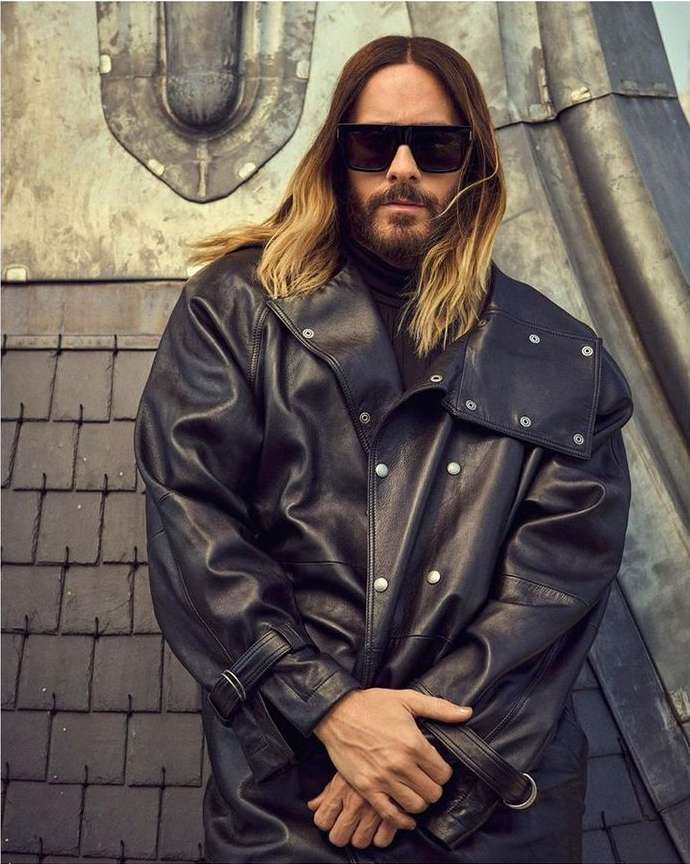 Adult male with long hair and sunglasses poses in a black leather jacket against a textured industrial wall, creating a fashion-forward portrait.
