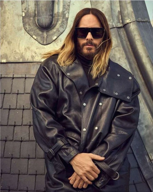 Adult male with long hair and sunglasses poses in a black leather jacket against a textured industrial wall, creating a fashion-forward portrait.