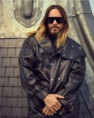 Adult male with long hair and sunglasses poses in a black leather jacket against a textured industrial wall, creating a fashion-forward portrait.
