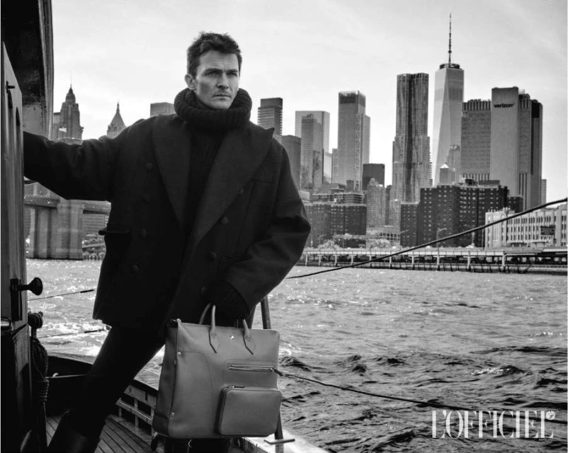 Monochrome shot of a man in a coat holding a briefcase on a boat, with the New York City skyline visible in the background.