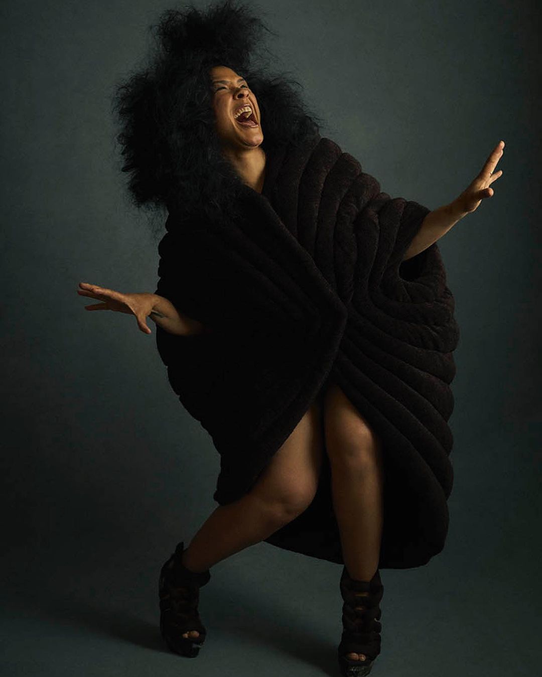 A joyful woman with an afro and dark draped clothing poses dramatically with arms outstretched, smiling against a dark background in a studio.