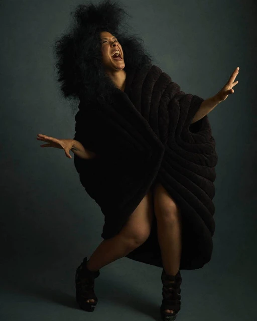 A joyful woman with an afro and dark draped clothing poses dramatically with arms outstretched, smiling against a dark background in a studio.