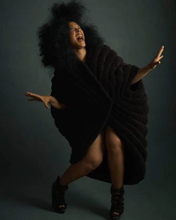 A joyful woman with an afro and dark draped clothing poses dramatically with arms outstretched, smiling against a dark background in a studio.