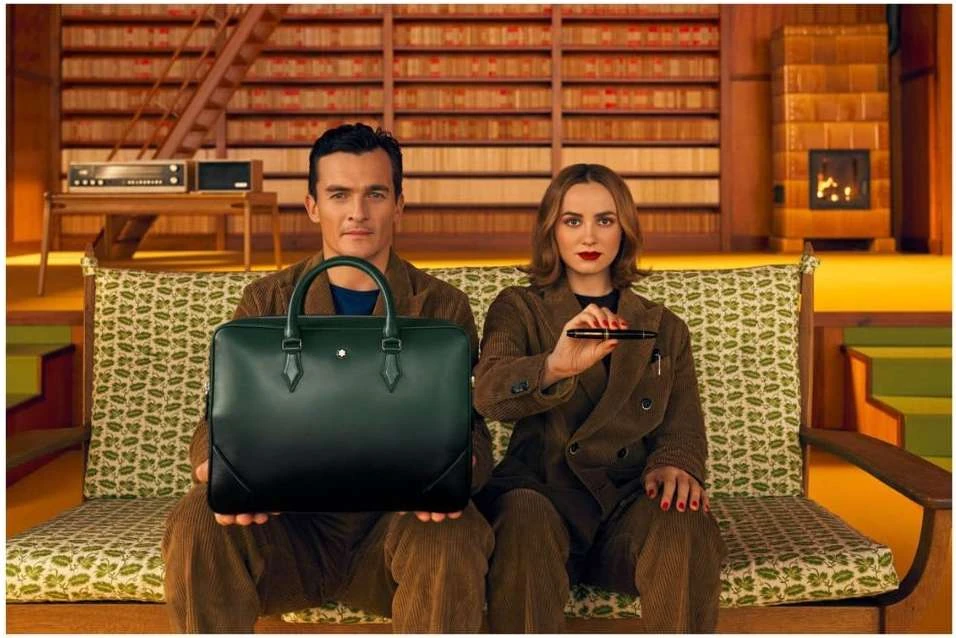 Stylish couple poses in a library-like setting, man holding a dark green leather bag, woman holding a pen, both in retro-style corduroy suits.