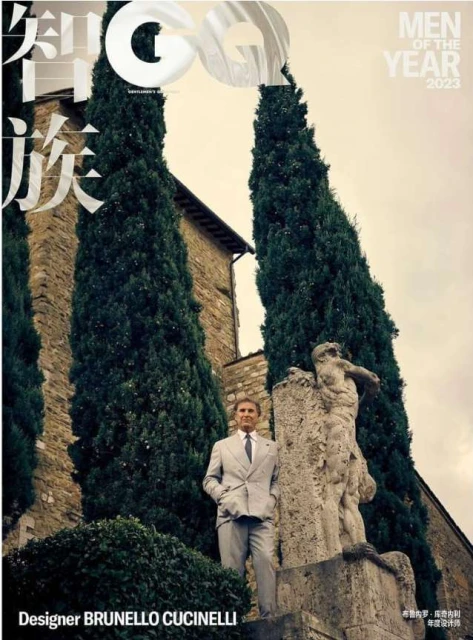 Brunello Cucinelli poses beside a classical statue in a light-grey suit for a magazine cover framed by tall trees and stone architecture.