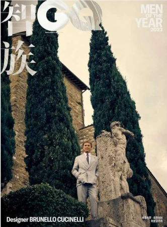 Brunello Cucinelli poses beside a classical statue in a light-grey suit for a magazine cover framed by tall trees and stone architecture.