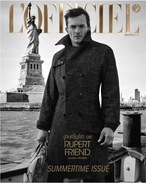 Magazine cover featuring Rupert Friend in a coat, holding a bag, with the Statue of Liberty in the background, and text overlay.