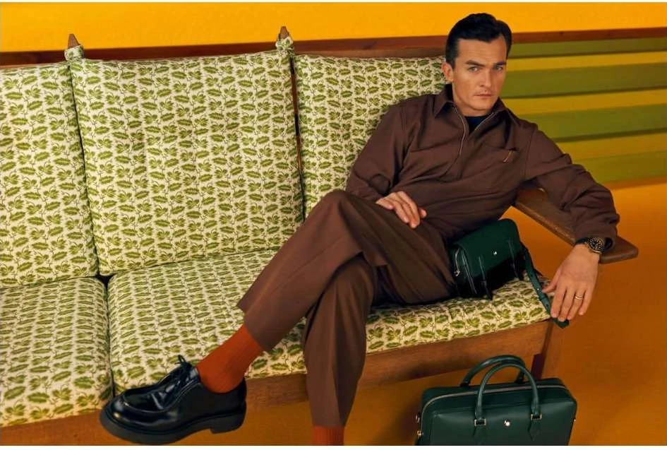 A man in a brown jumpsuit poses on a green floral couch with matching green bag and briefcase against a retro yellow background.