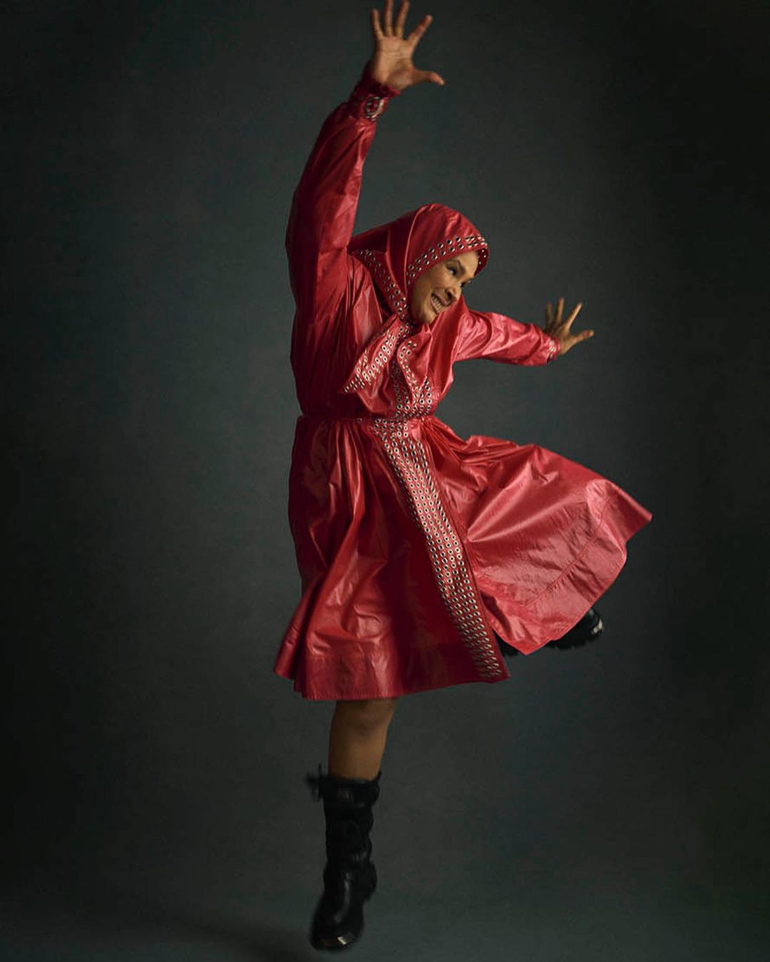 A person in a red hooded dress leaps mid-air against a dark grey background, arms raised and legs bent, striking a joyful, dynamic pose.
