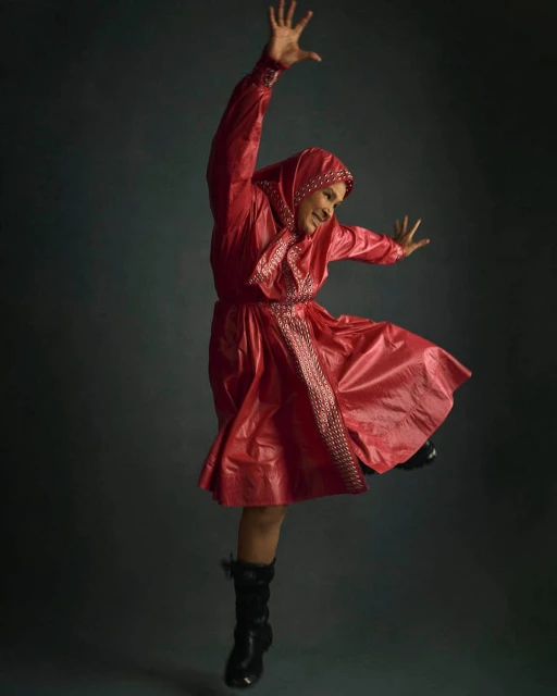 A person in a red hooded dress leaps mid-air against a dark grey background, arms raised and legs bent, striking a joyful, dynamic pose.