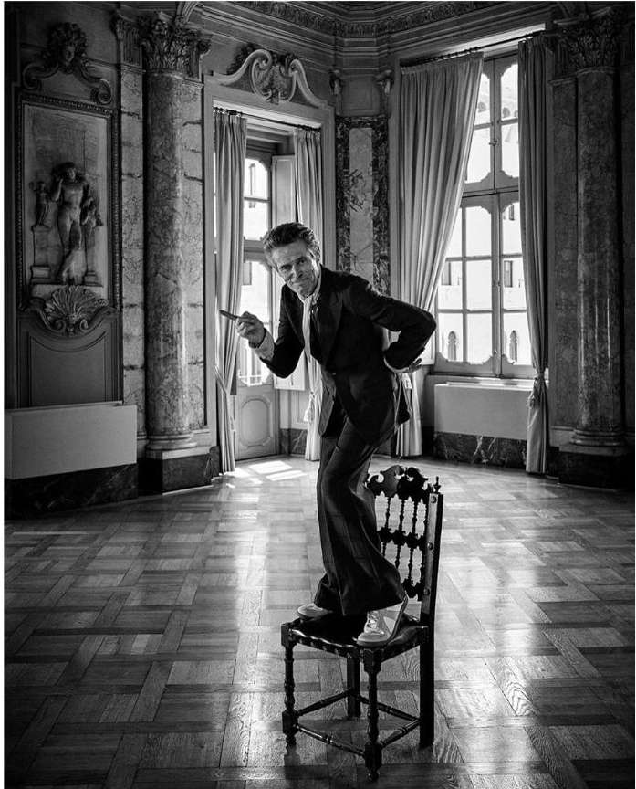 Black and white portrait of an adult male standing on a chair in a tuxedo, in an ornate room with herringbone floors and large windows.