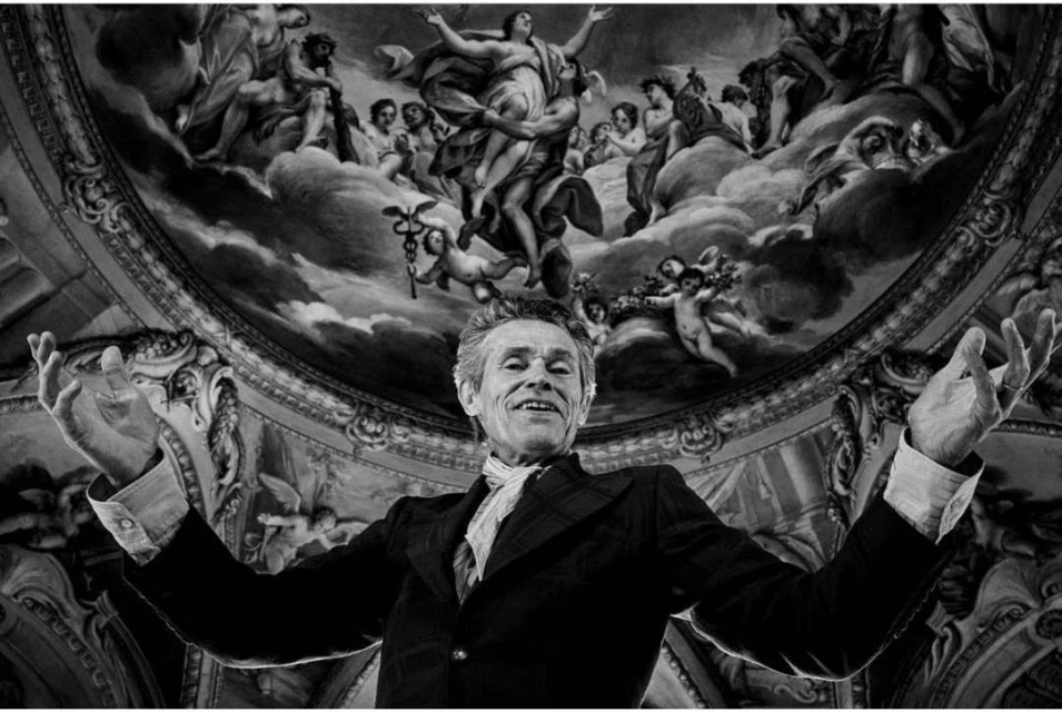 Black-and-white portrait of an adult man with outstretched arms, set against a backdrop of a historical ceiling mural with figures.