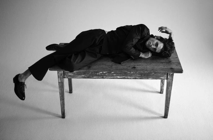 Monochrome portrait of a man reclining on a weathered wooden table wearing a dark suit and dress shoes against a plain background.