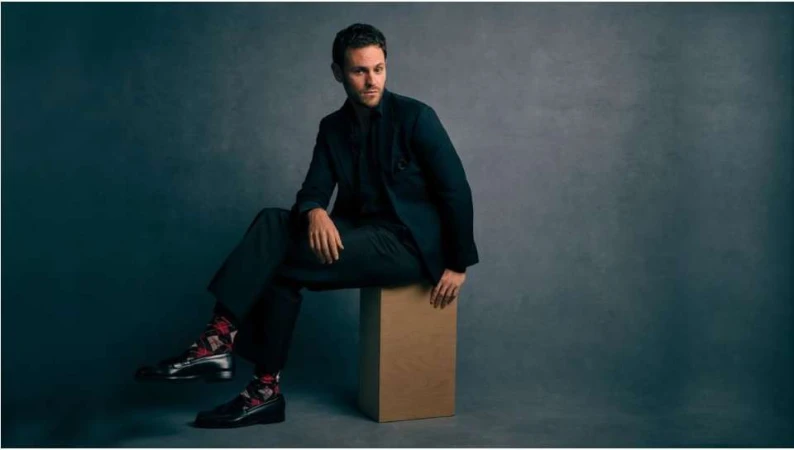 Man in a dark suit seated on a wooden box against a textured grey studio backdrop, photographed by Charlie Gray.