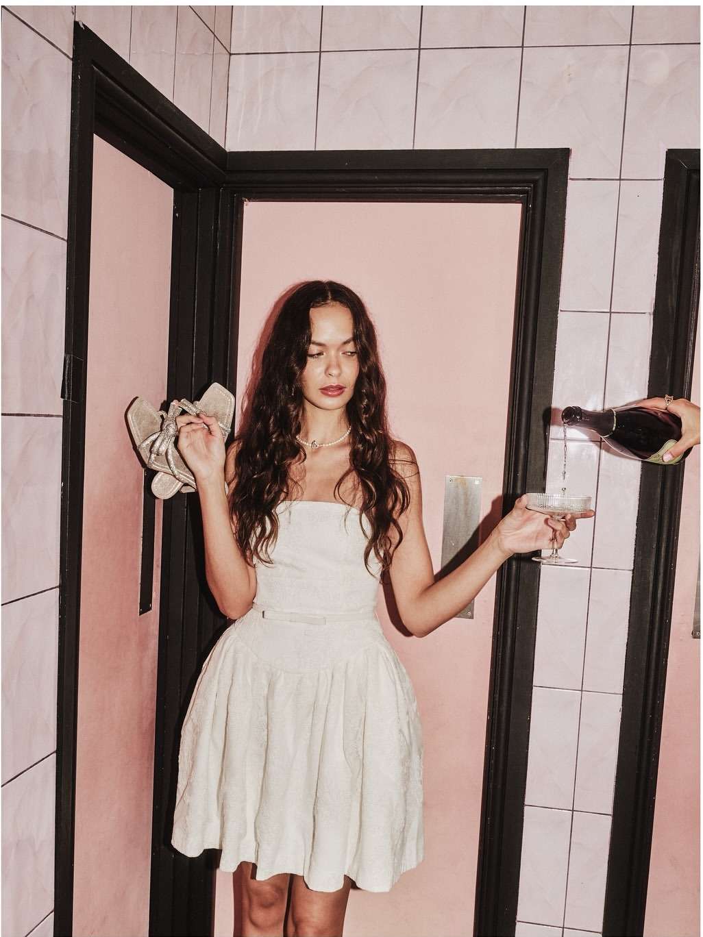 Elegant woman in short dress poses in tiled bathroom as champagne is poured into glass. Fashion and celebration concept.
